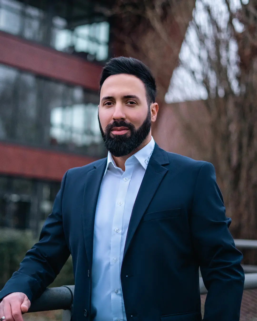 Mann mit Bart in einem Anzug, der an einem Gel&auml;nder steht und in die Kamera schaut.