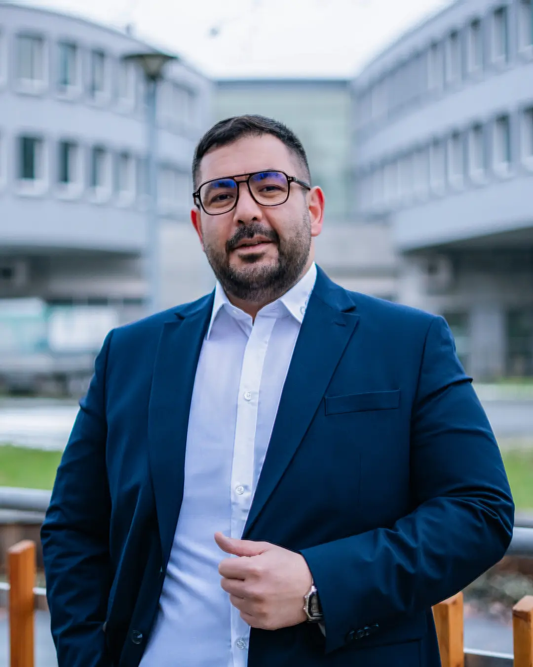 Mann in einem Anzug mit Brille steht vor modernen Geb&auml;uden und l&auml;chelt leicht.